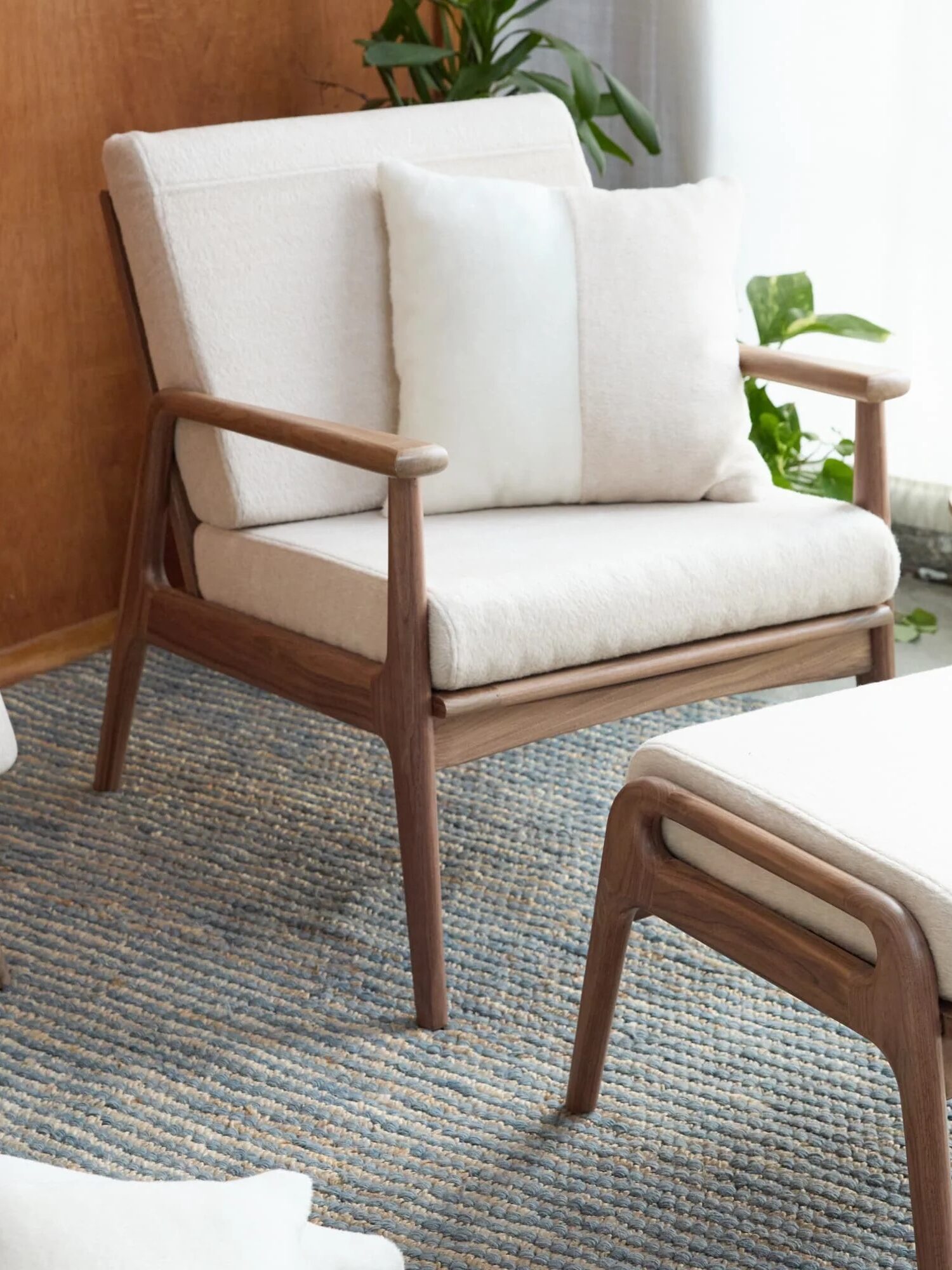 A wooden chair and matching ottoman with white cushions sit on a woven rug next to a small round side table holding a glass, near a window with white curtains and a wooden wall.