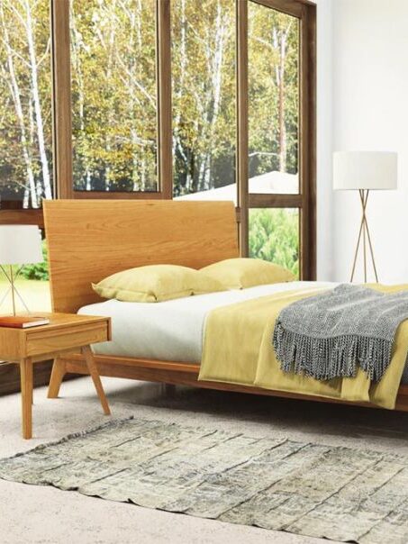 A modern bedroom with large windows, a wooden bed with yellow and gray bedding, a matching nightstand, a dresser with a triple mirror, a floor lamp, and a rug on a light-colored floor.