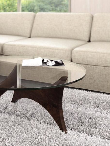 A round glass coffee table with a dark wooden base is placed on a light gray rug in front of a beige sectional sofa. A few items are on the table.