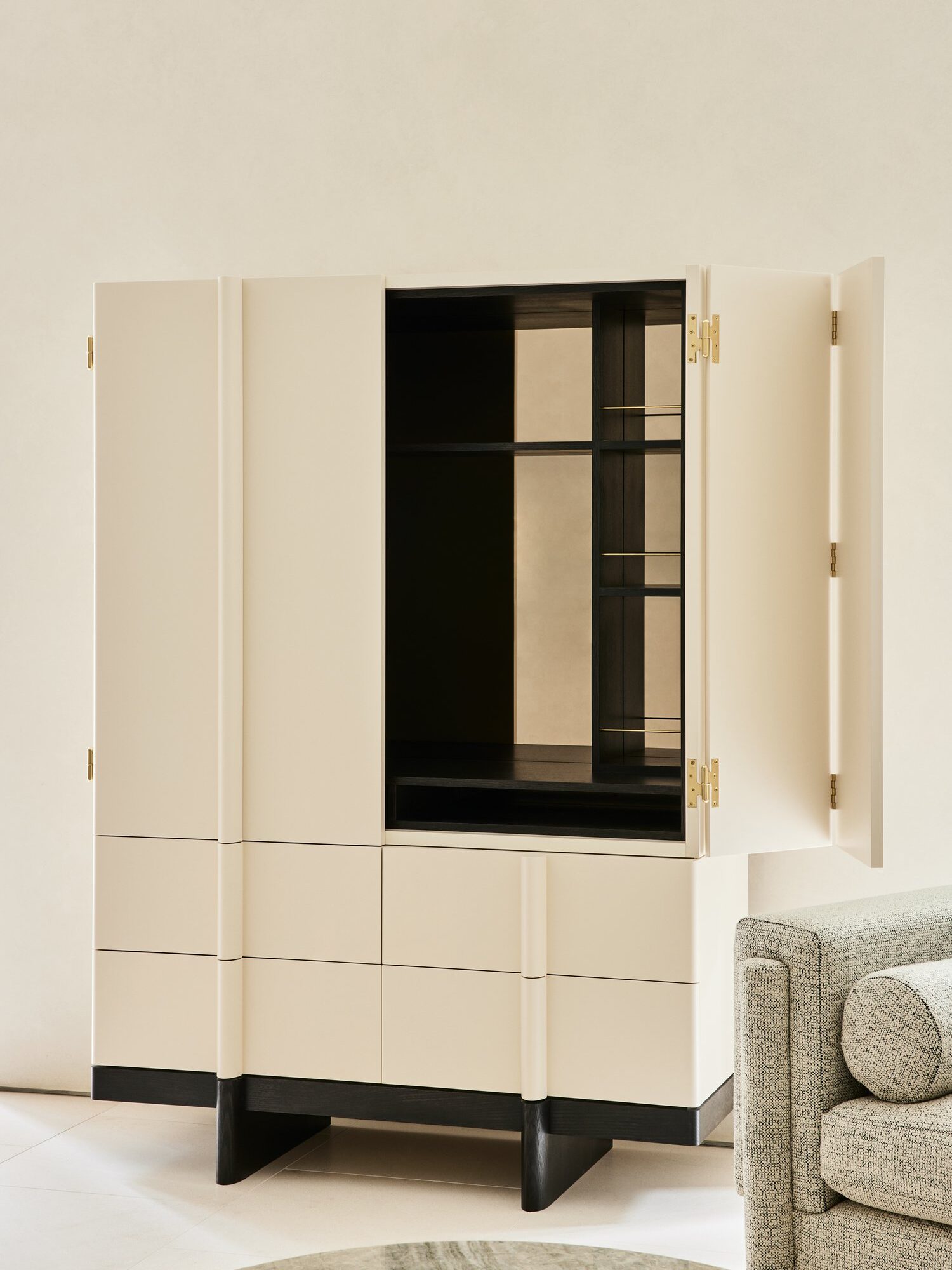 A beige modern cabinet with gold hinges and open doors revealing black interior shelves. It stands next to a grey upholstered sofa in a neutral-toned room.