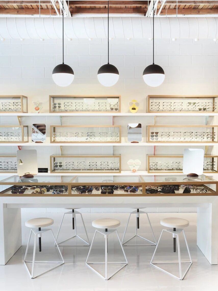 A modern eyewear store with glasses displayed on shelves and a central display table. Three stools are placed around the table. The interior features a mix of white and wood accents and potted plants.