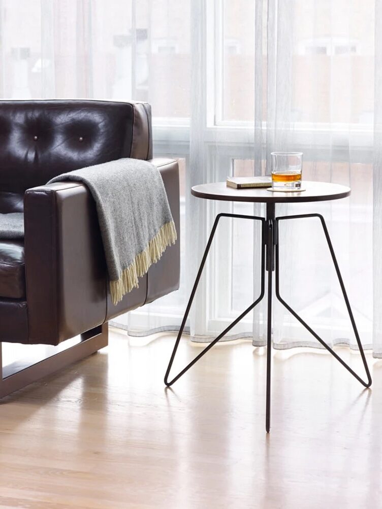 A brown leather sofa with a gray throw is next to a small round side table with a glass of whiskey and a book on it, set in front of a large window with translucent curtains.