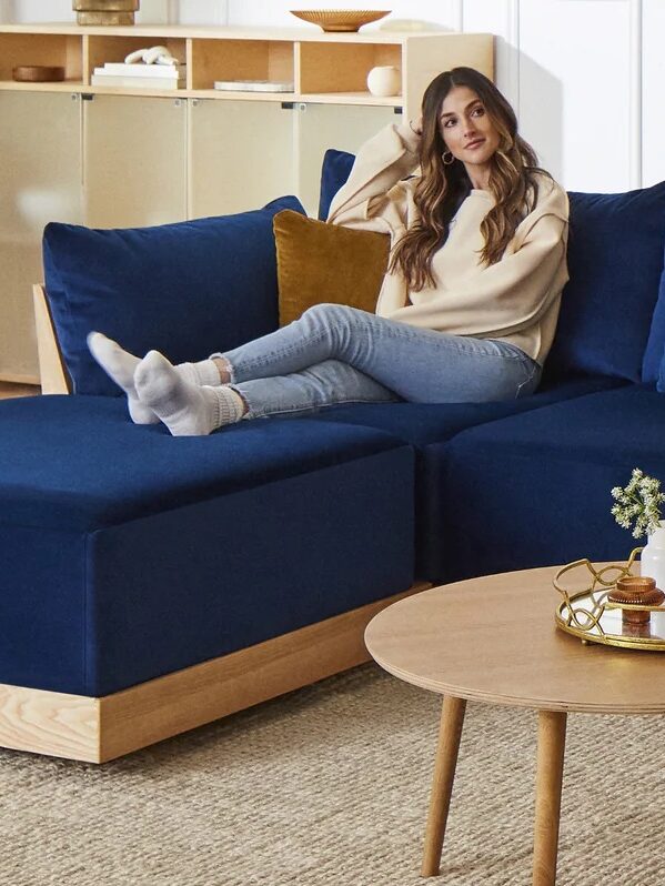 A woman sits on a blue sectional sofa with mustard cushions in a modern living room featuring white walls, a wooden coffee table, a floor lamp, and shelves.