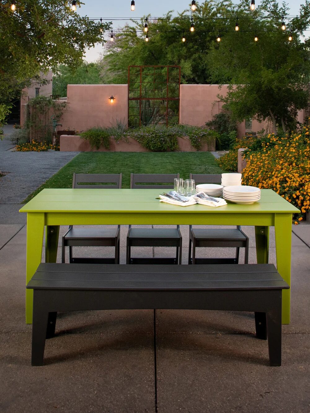 Outdoor dining area with a lime green table, black bench, and three chairs set in a garden patio. String lights hang above, and the table holds stacked plates and glasses. Bushes and greenery surround the setup.