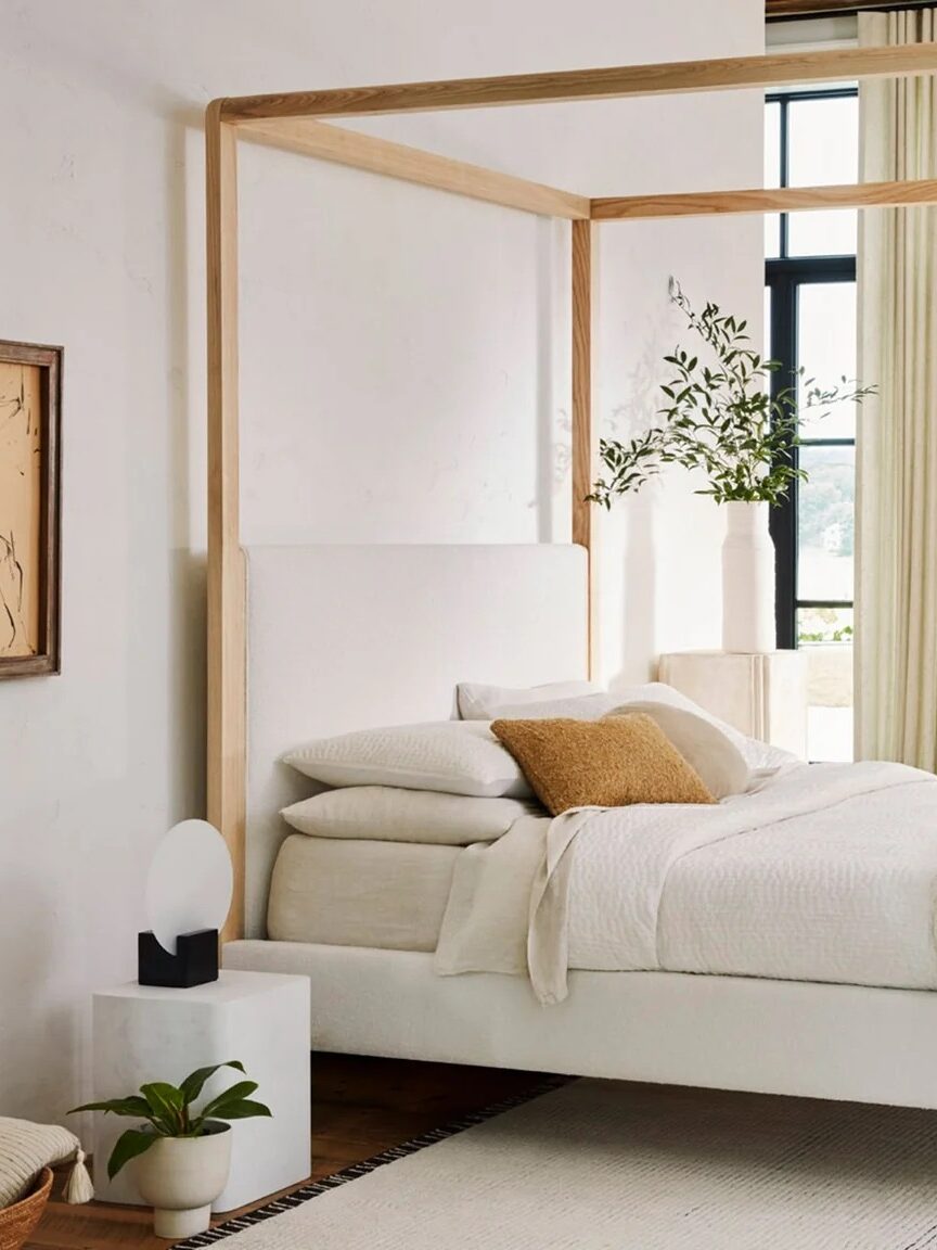 A modern bedroom with a four-poster bed, neutral bedding, a green ottoman, white armchair, and large windows with beige curtains. The room features minimalistic decor and wooden accents.