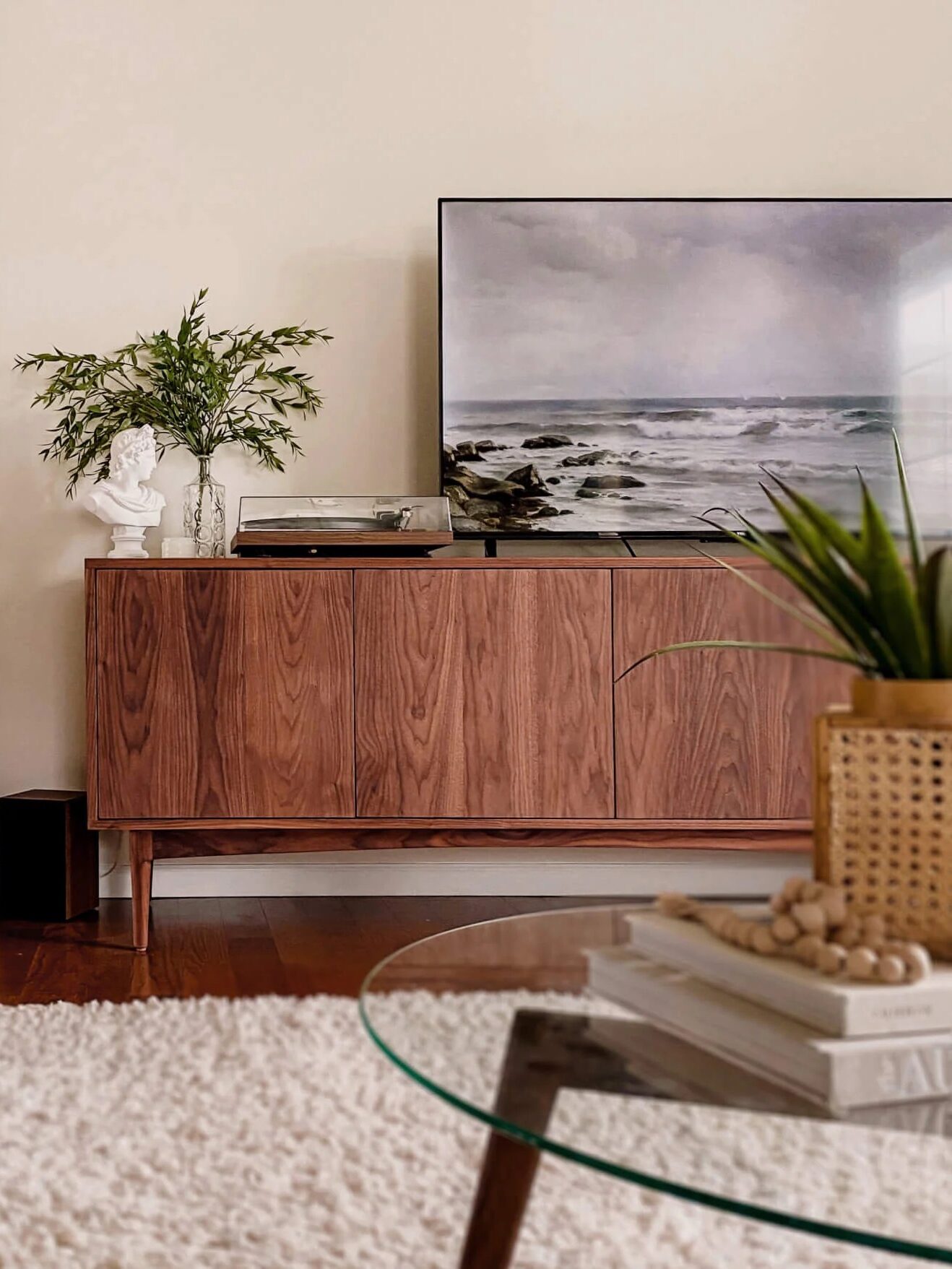 A modern living room features a wooden sideboard with a TV displaying a seascape, a plant, and a bust sculpture. A large mirror reflects an armchair, and a glass coffee table sits on a white rug.
