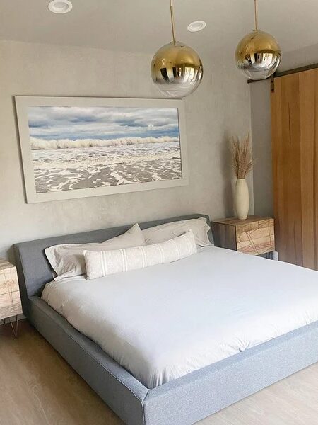 Modern bedroom with a low gray bed, wooden sliding door, nightstands, and hanging light fixtures. Artwork depicting a beach scene is mounted on the wall. Adjoining bathroom visible through the open door.