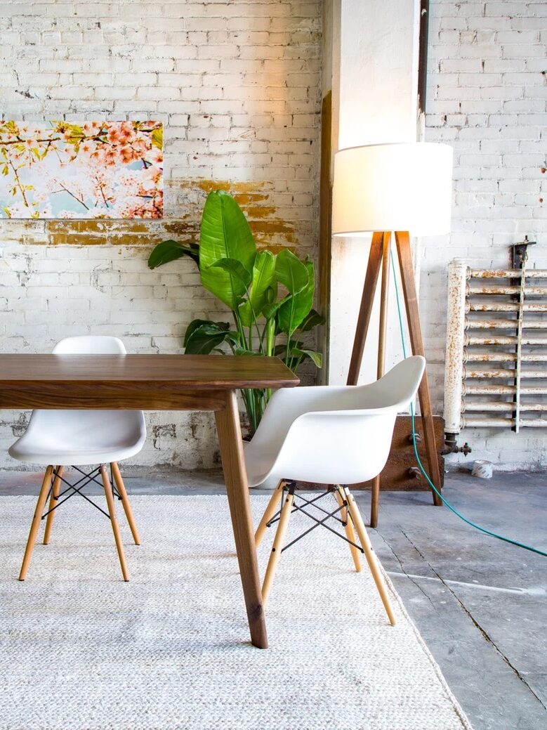 A modern dining area features a wooden table, four white chairs with wooden legs, tall indoor plants, a floor lamp, and a colorful wall painting against a white brick wall background.