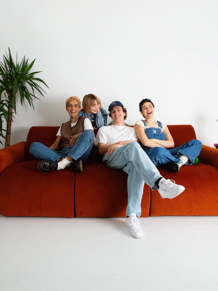 Four people sitting together on an orange sofa in a white room. A lamp and a plant are visible on either side of the sofa.