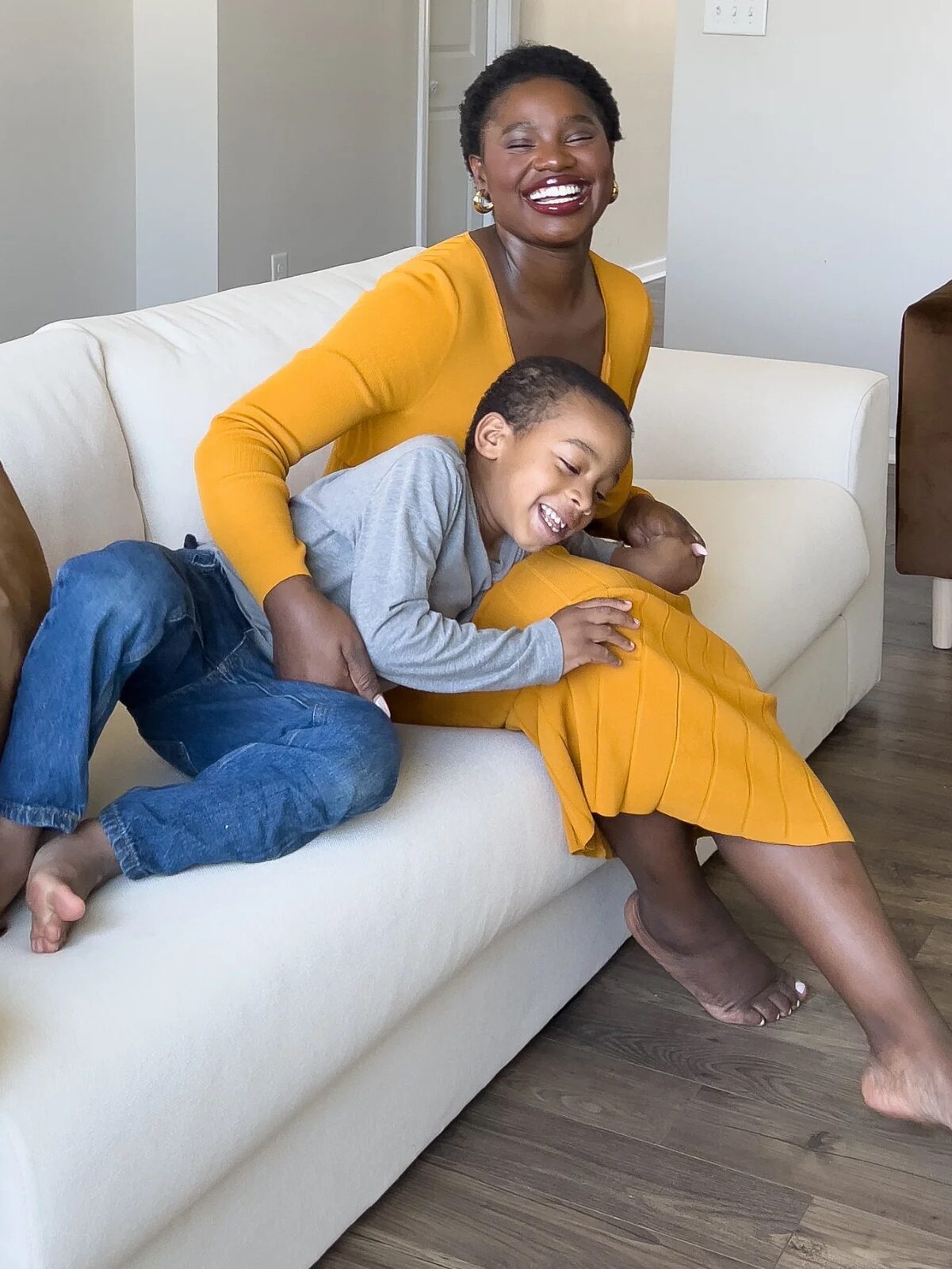 A woman in a yellow dress sits on a white couch, smiling and embracing a young boy who is laughing and leaning against her.