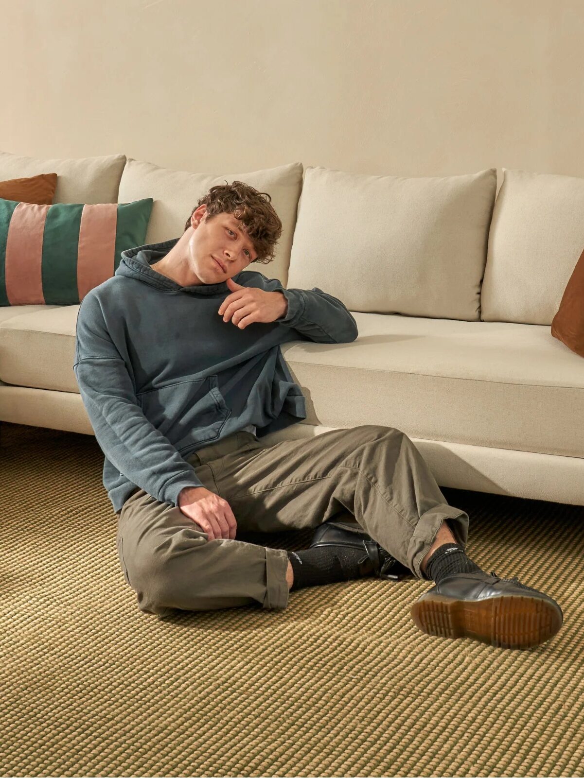 A person wearing a blue hoodie and khaki pants sits on the floor resting against a beige sofa in a living room with a beige rug, pillows, a side table with books, and a vase with flowers.