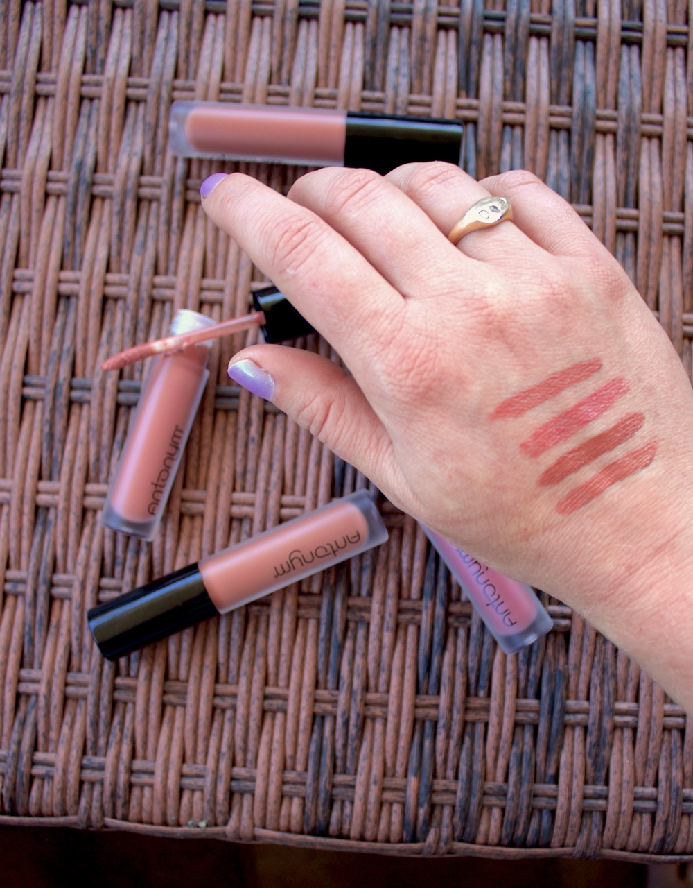 A hand with five swatches of different shades of lipstick on the back, next to five lipstick tubes placed on a wicker surface.