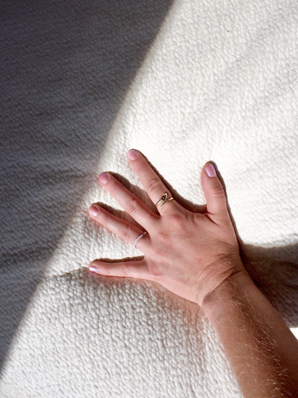 A hand with a gold ring on the middle finger and a silver ring on the ring finger rests on an organic mattress's textured white surface, illuminated by sunlight.