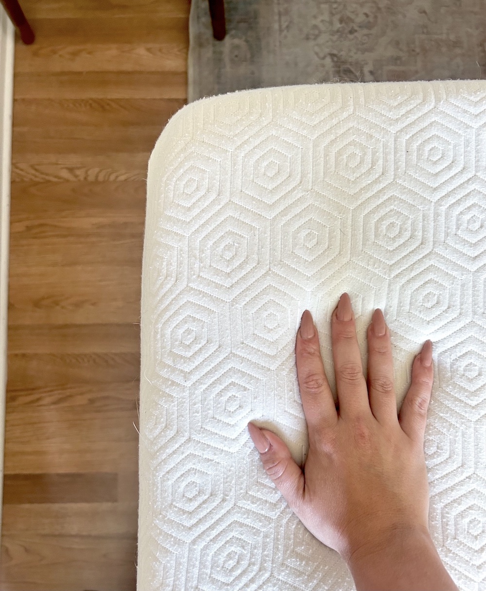 A hand resting on a white, organic mattress cover with a hexagonal pattern. The background includes a wooden floor and part of a rug.