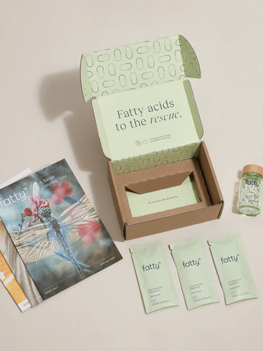 Open box with the text "Fatty acids to the rescue" containing three small green sachets labeled "fatty," a small glass jar with a wooden lid, and a magazine featuring a dragonfly on the cover.