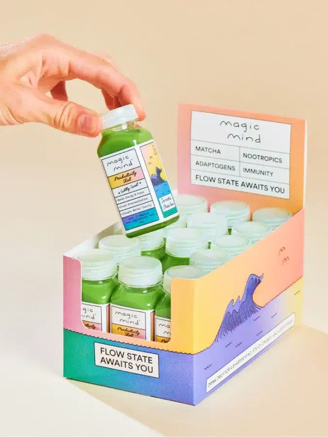 Hand holding a small green bottle from an open box labeled "magic mind," which contains multiple similar bottles. The box promotes benefits like matcha, nootropics, adaptogens, and immunity support.