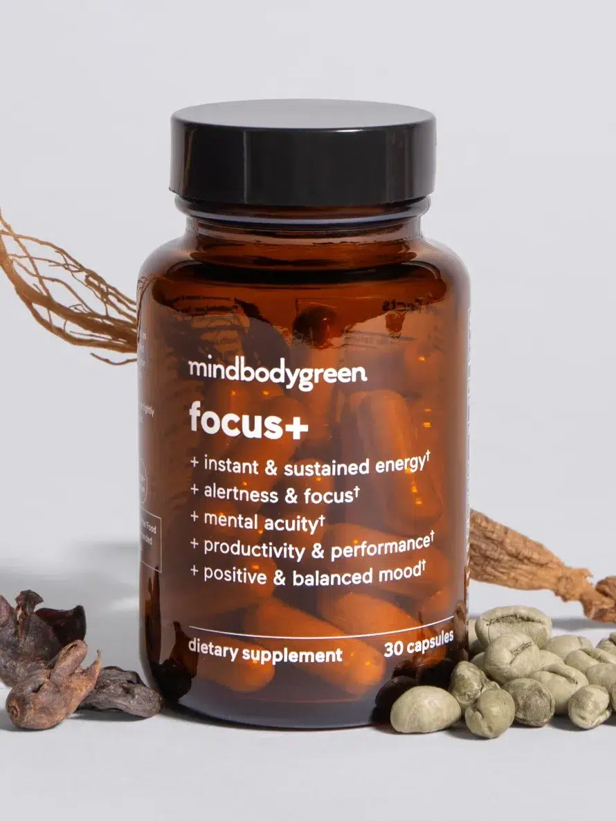 A bottle of mindbodygreen focus+ dietary supplement with 30 capsules, surrounded by various herbal ingredients. The label highlights benefits like energy, alertness, mental acuity, productivity, and mood balance.