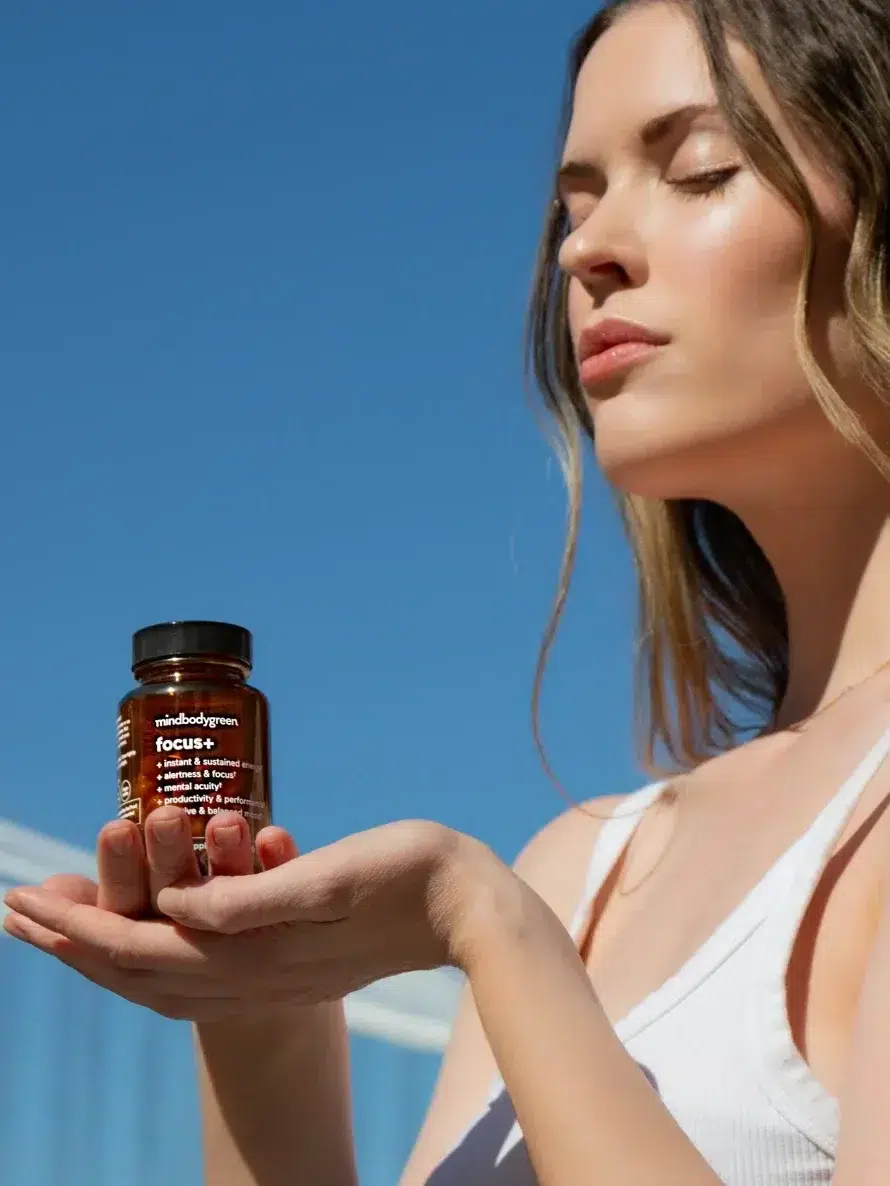 A woman in a white top holds a small bottle labeled "focus" with both hands against a blue sky background, eyes closed.