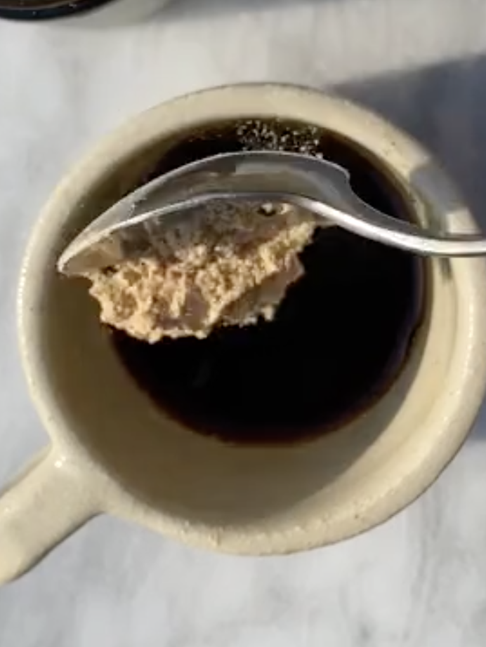 A spoon holding a chunk of beige substance is held over a cup of dark liquid on a white surface.