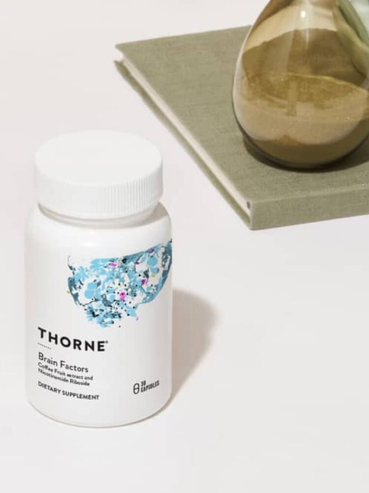 A bottle of Thorne Brain Factors dietary supplement sits on a white surface next to a green-gray book and a sand hourglass.