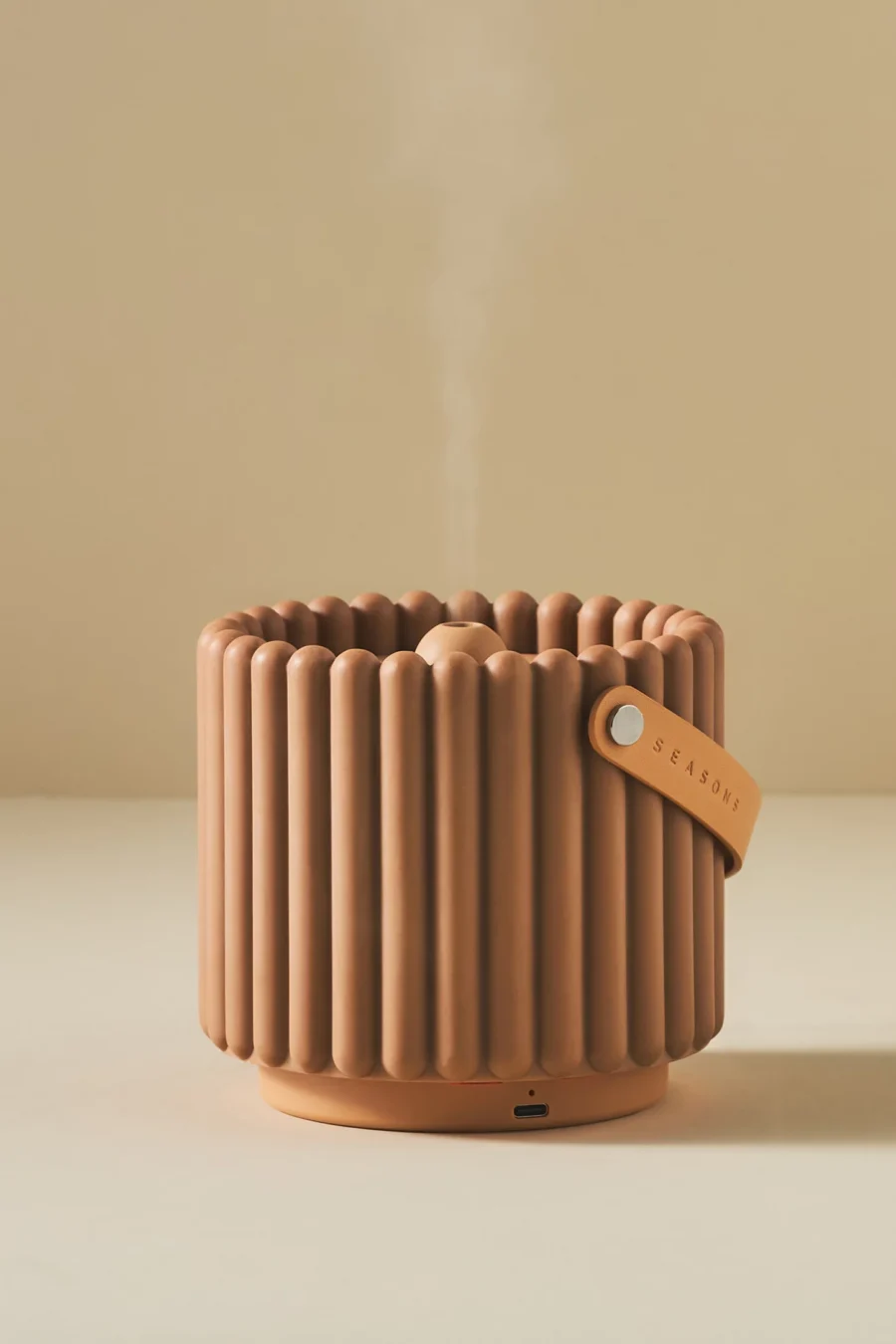 A ribbed, cylindrical humidifier emitting steam, with a leather strap labeled "Seasons" and a USB-C port at the base, placed on a beige surface.