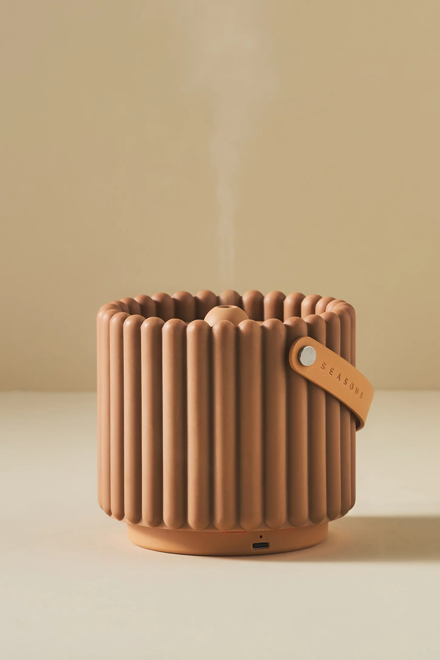 A ribbed, cylindrical humidifier emitting steam, with a leather strap labeled "Seasons" and a USB-C port at the base, placed on a beige surface.