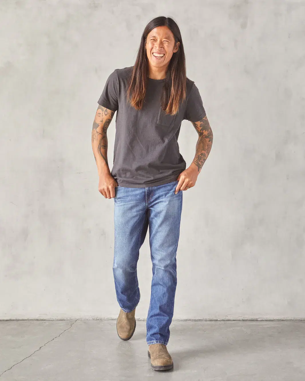 A person with long hair, wearing a dark t-shirt, blue jeans, and brown shoes, is smiling while walking towards the camera against a gray background.