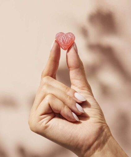 A hand with light-colored nail polish holds a small, pink heart-shaped candy with a "V" on it.