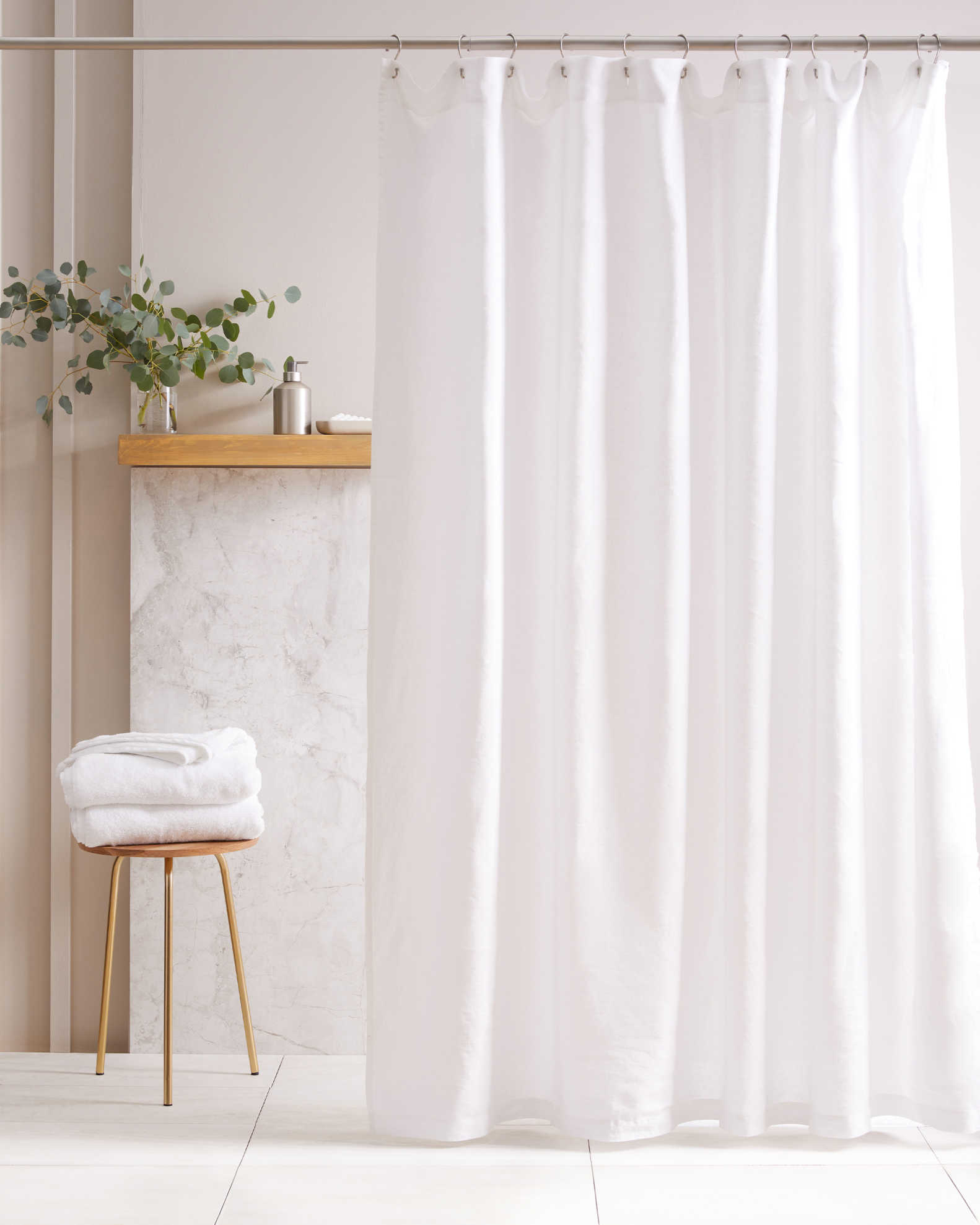 A white shower curtain hangs in a bathroom with a towel on a stool, a small shelf with a plant, and toiletries in the background.