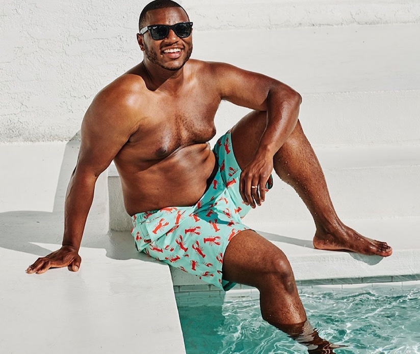 A man in shorts and sunglasses sits on white steps by a swimming pool, smiling.