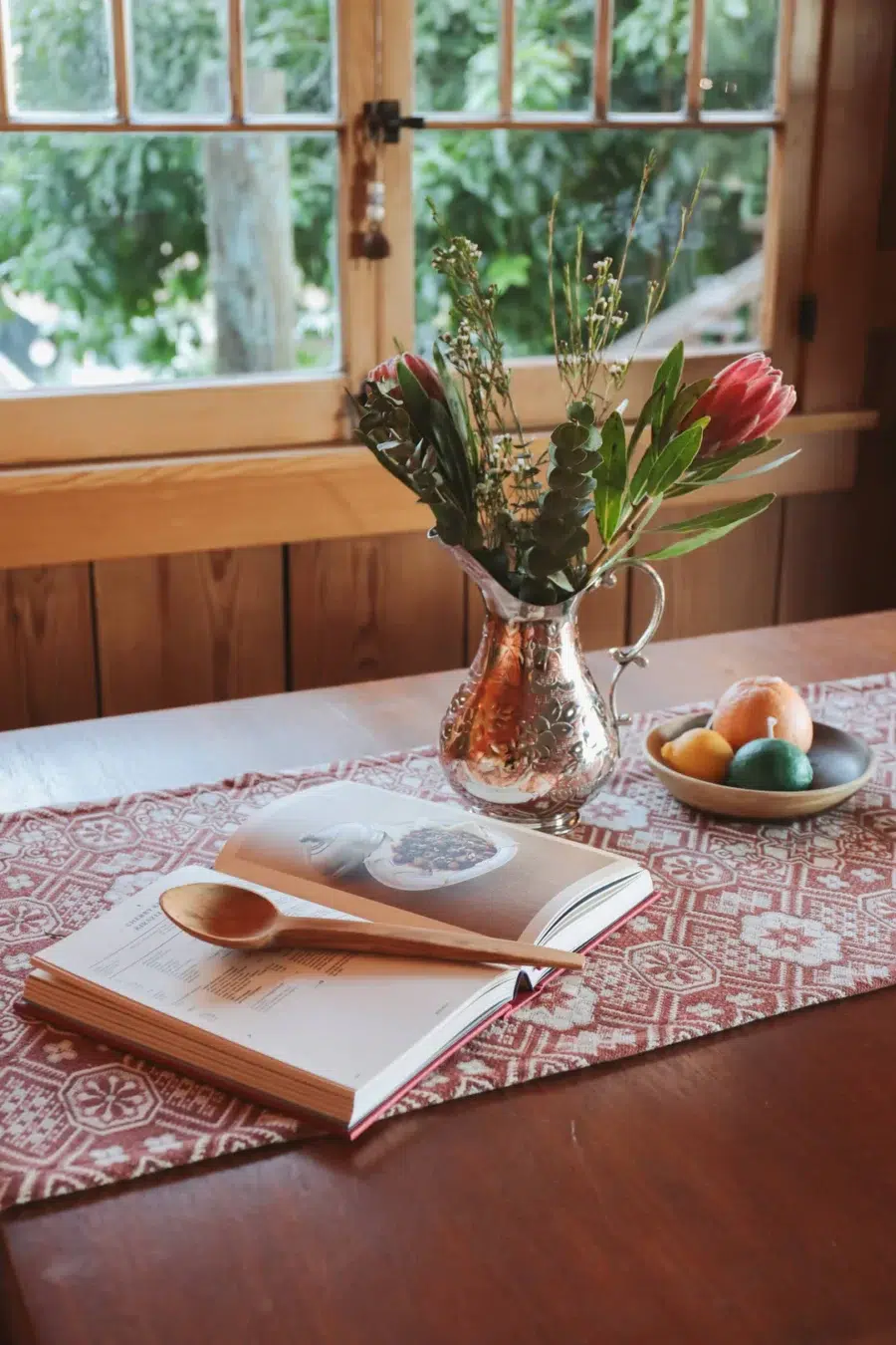 A wooden table with an open cookbook, wooden spoon, vase with flowers, a bowl of fruits, and a window with greenery outside.