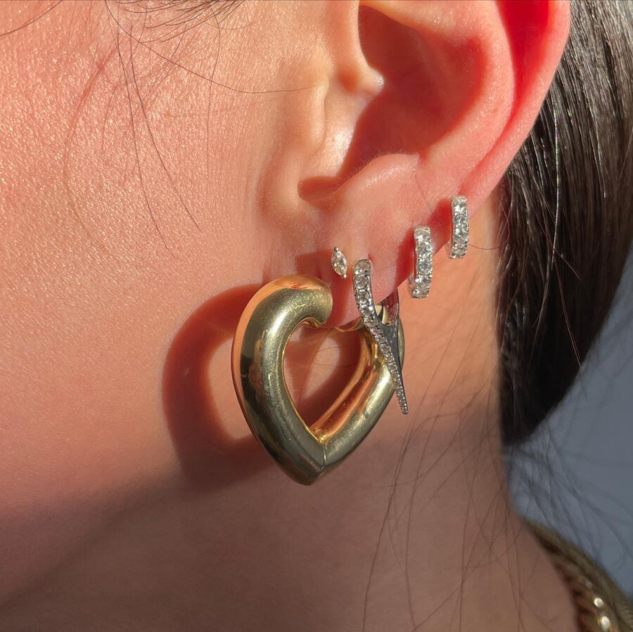 Close-up of a person's ear wearing multiple earrings, including a large gold heart-shaped hoop and several small, sparkling, silver studs and hoops.