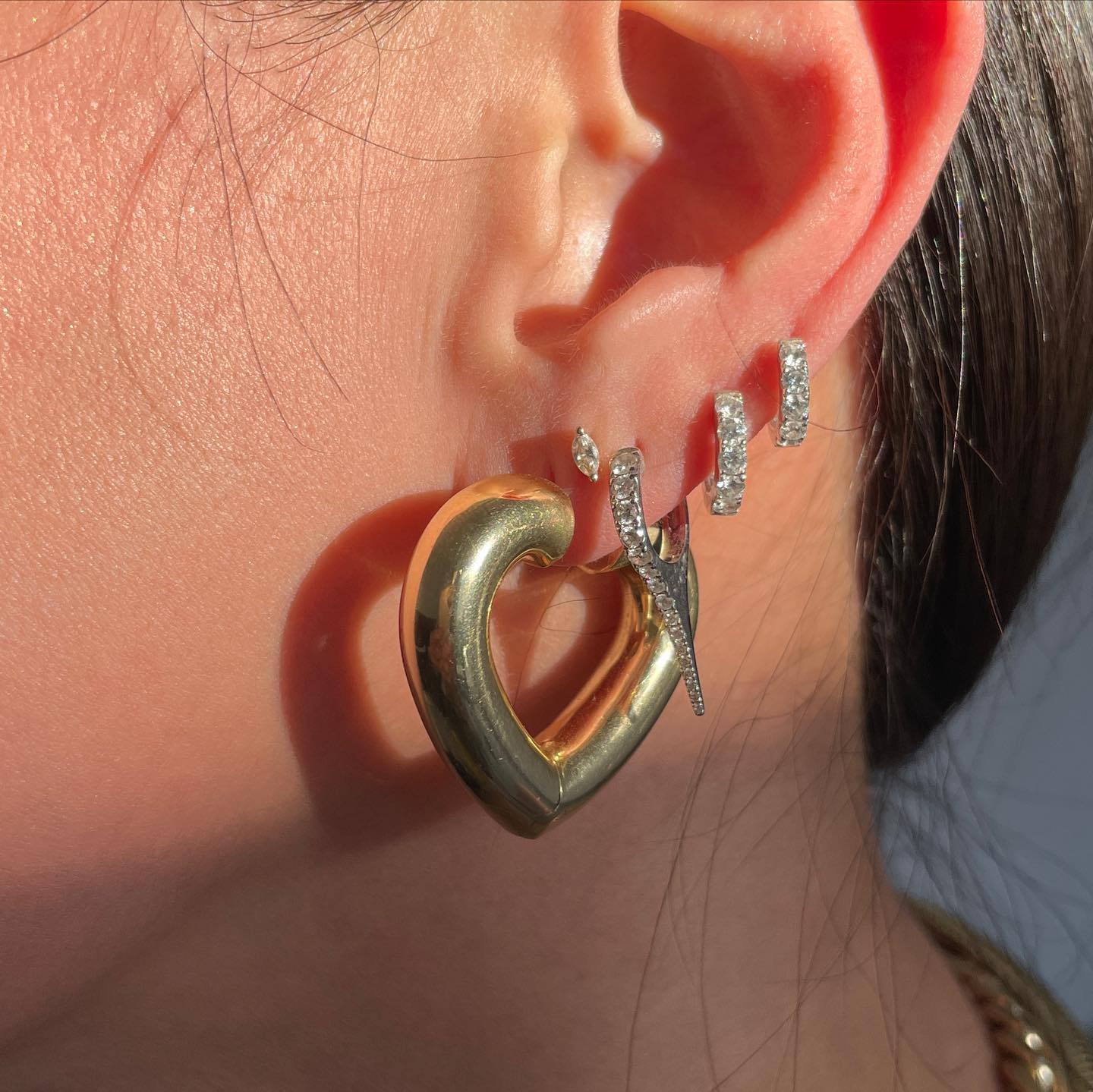 Close-up of a person's ear wearing multiple earrings, including a large gold heart-shaped hoop and several small, sparkling, silver studs and hoops.