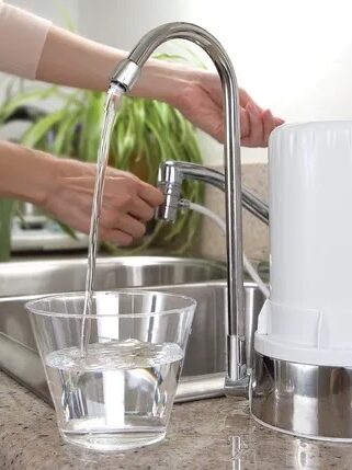 A person uses a water filter system at a kitchen sink, filling a glass with filtered water.