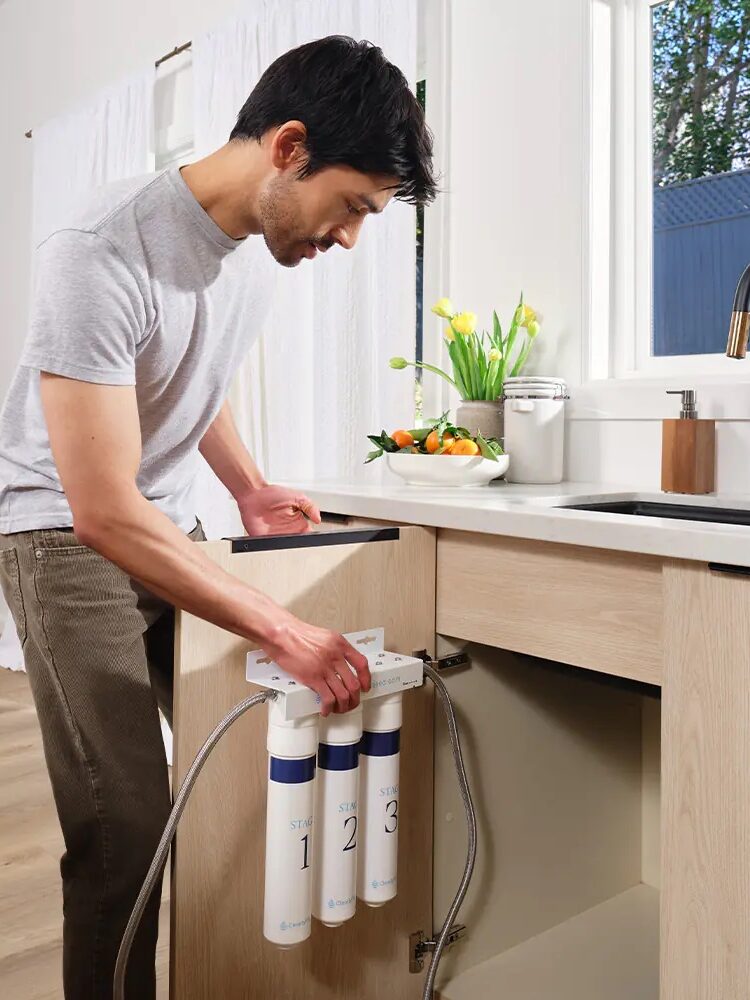 A person is installing a three-stage water filtration system under a kitchen sink. The kitchen has light wood cabinets, a black faucet, and a window with a view of trees outside.