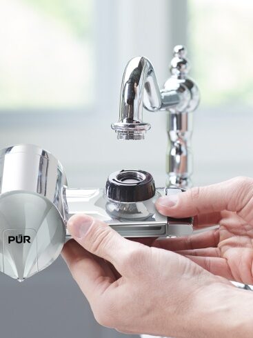 Hands installing a PUR water filter attachment to a kitchen faucet.