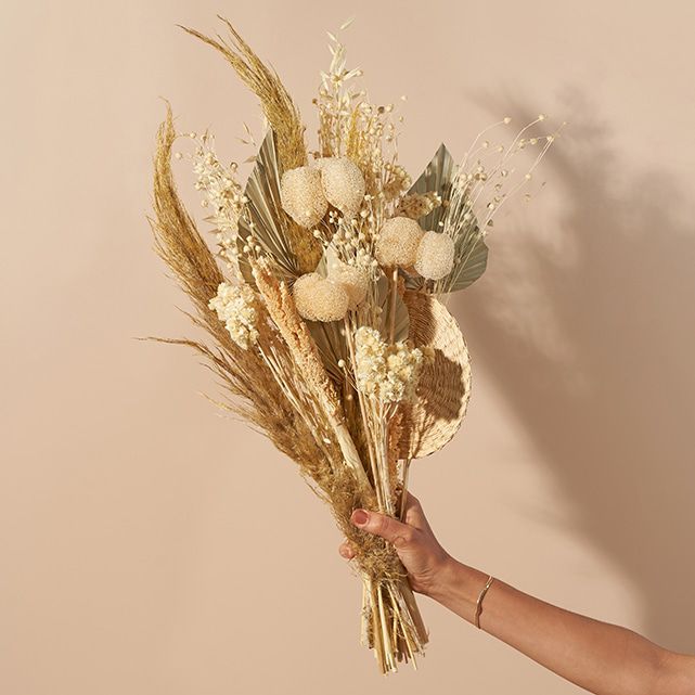 A hand holds a bouquet of dried flowers and grasses, including pampas grass, palm leaves, and various white, beige, and brown botanicals against a plain beige background.