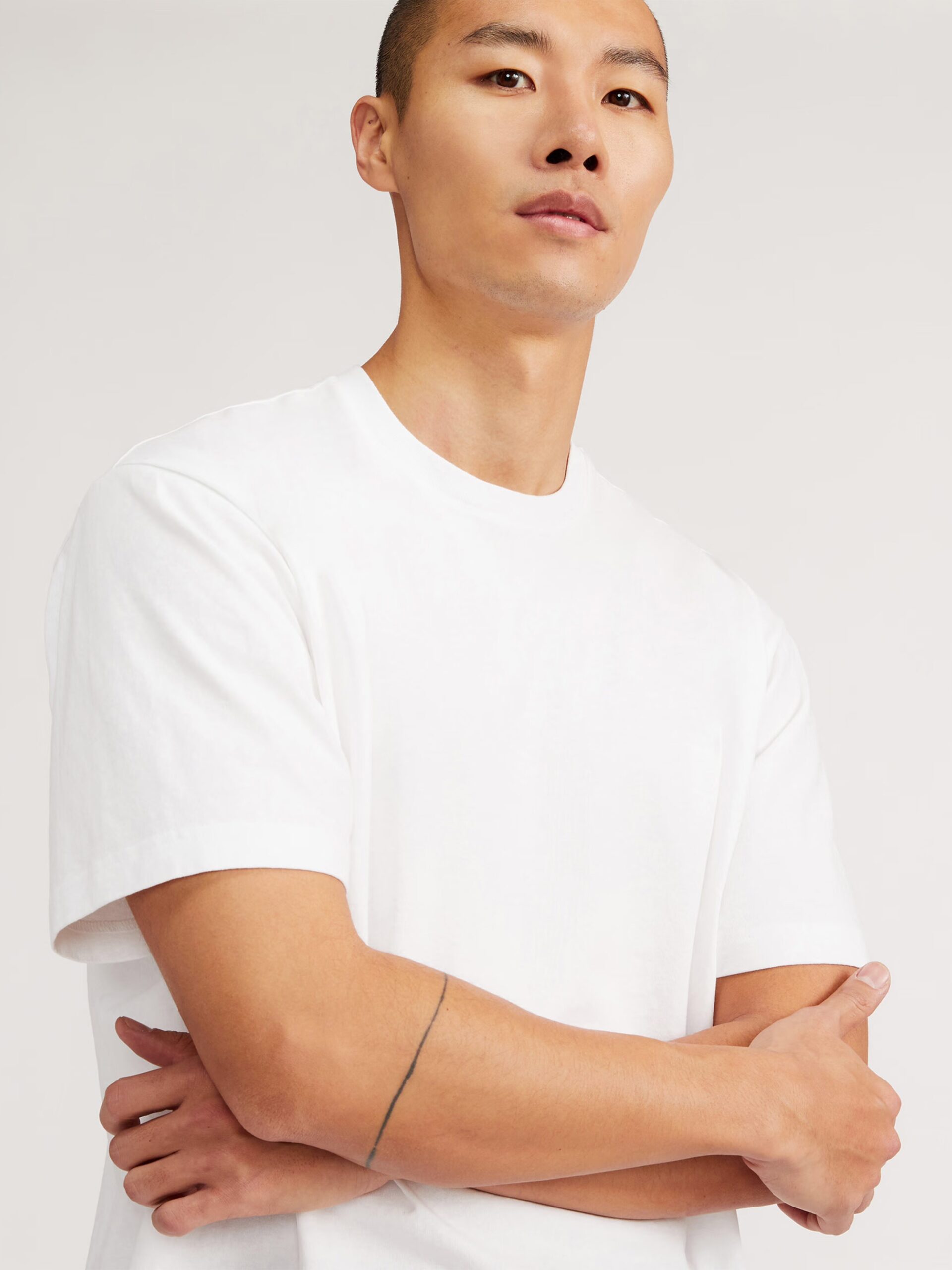 A person with a shaved head wearing a plain white T-shirt stands with their arms crossed against a neutral background.