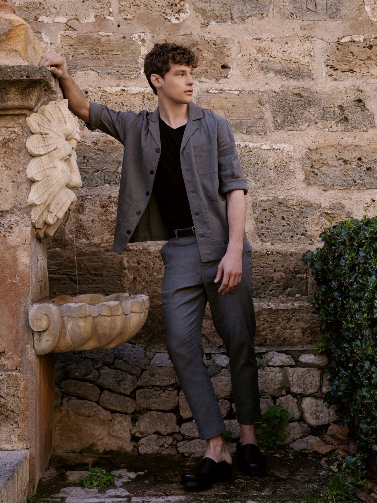 A man wearing a gray suit and black shirt stands by an ancient stone fountain, resting one hand on its edge. The background consists of a weathered stone wall and some greenery.