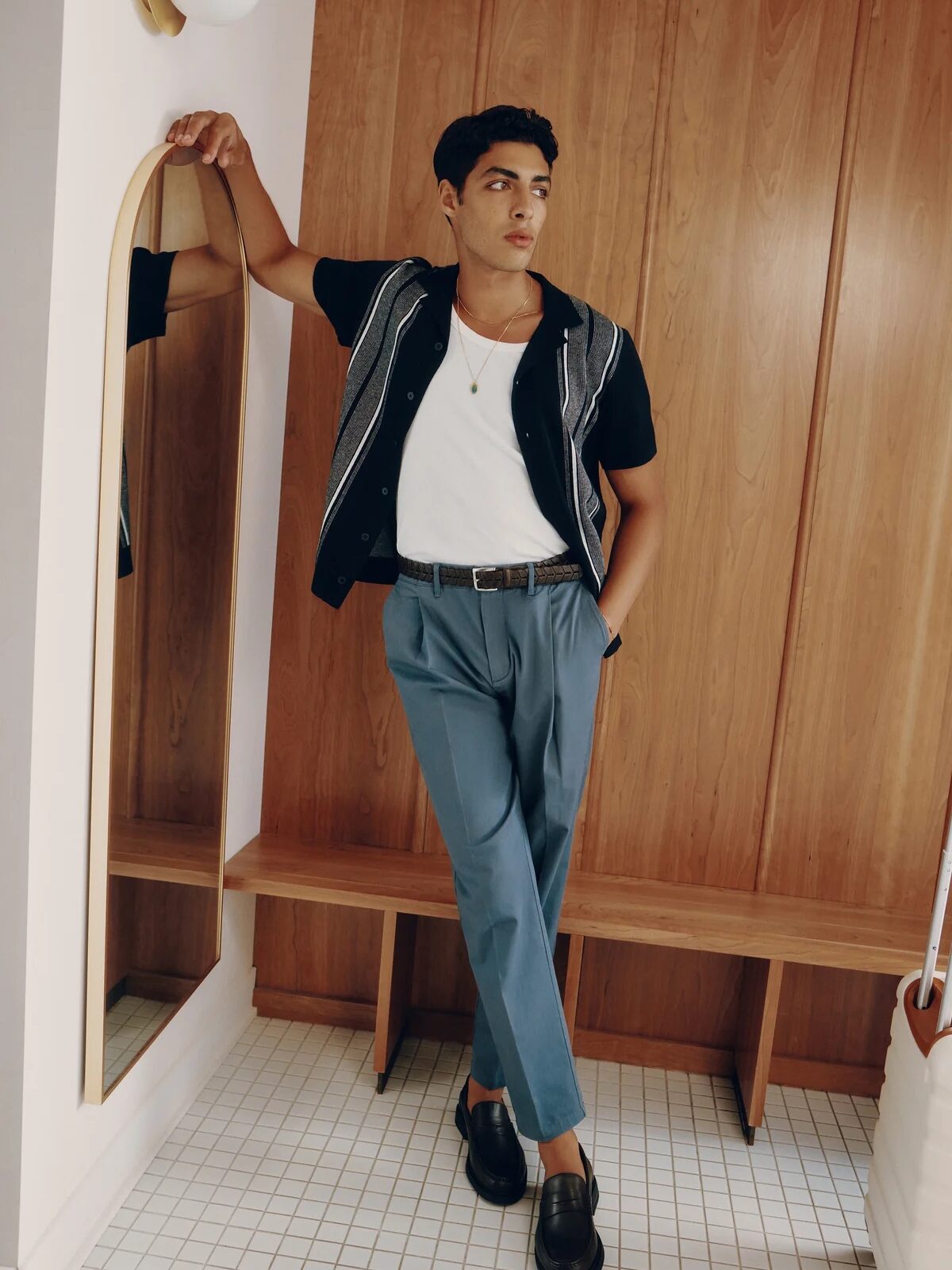 A model wearing an open, button down collared shirt in black and grey, with a white tank underneath and blue trousers, from Frank & Oak. 