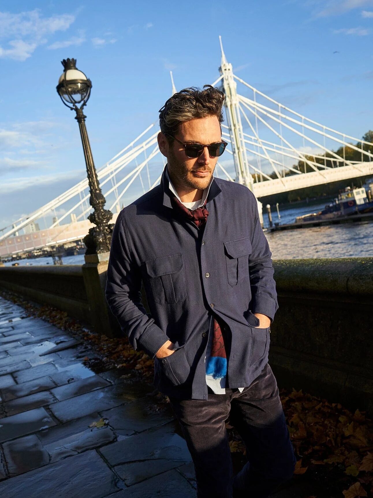 A man wearing sunglasses and a navy jacket walks along a riverside path with a lamp post and bridge in the background.