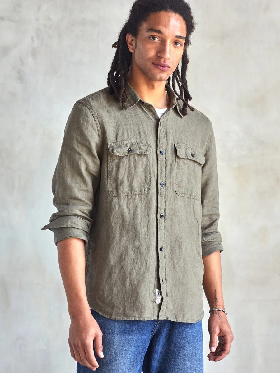 A person with long braided hair is wearing a green button-up shirt paired with blue jeans. They are standing against a plain, light-colored background.