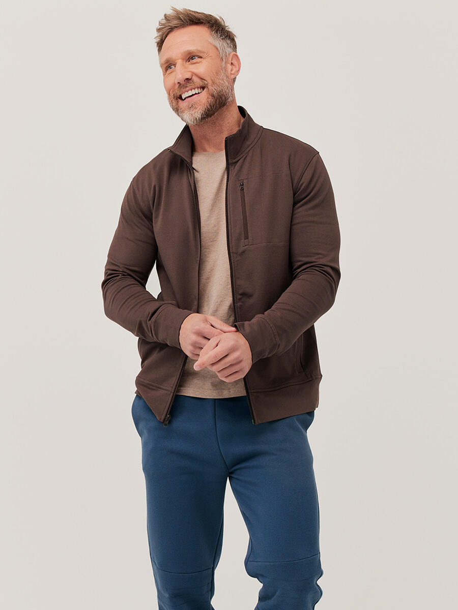 A man with short hair and a beard is standing against a plain background. He is wearing a brown zip-up jacket over a beige shirt and blue pants. He is smiling and has his hands together.