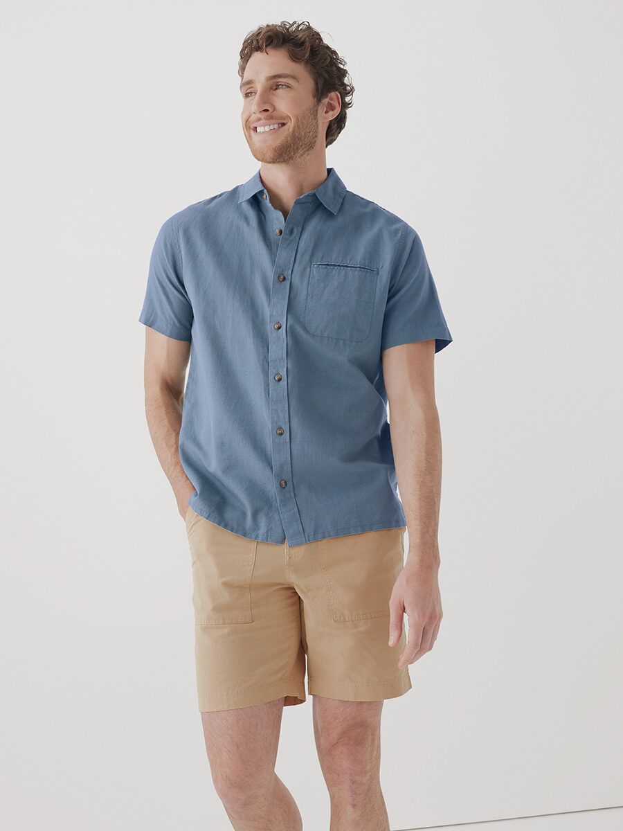 A man wearing a short-sleeve blue button-up shirt and beige shorts, standing and smiling against a plain white background.