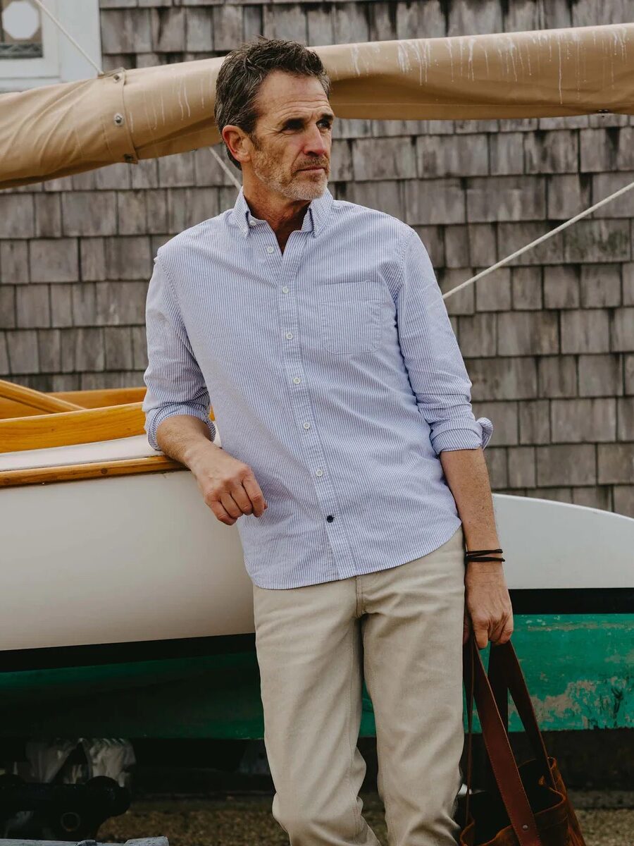 A man in a light blue button-up shirt and beige pants stands by a boat dock, holding a brown bag, with a wooden boat and building with wood shingles in the background.