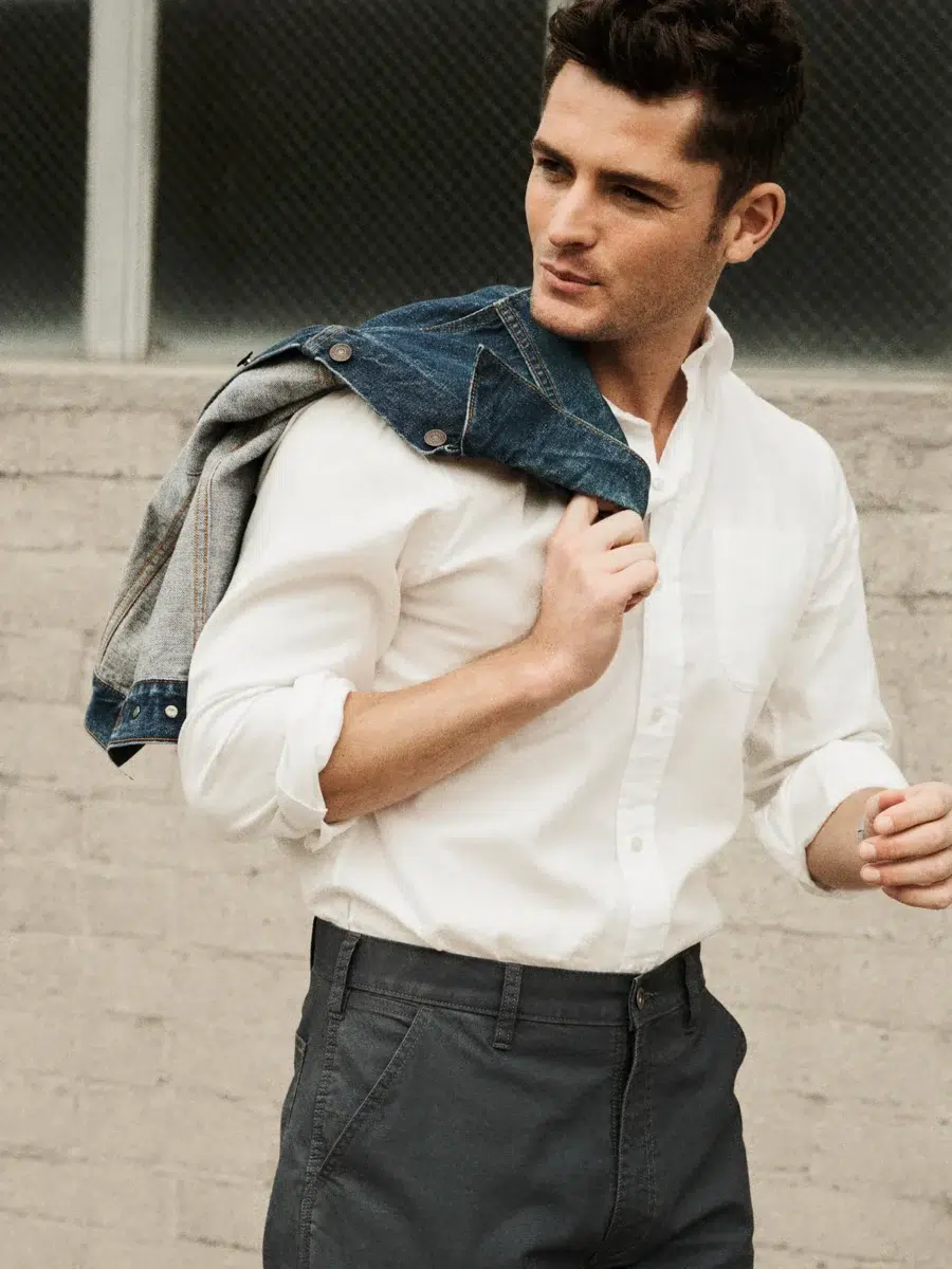 A man wearing a white shirt and grey pants holds a denim jacket over his shoulder while standing in front of a brick wall.
