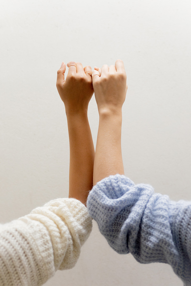 Two hands with interlocked pinky fingers, wearing pastel-colored sweaters, are raised against a plain, light background.