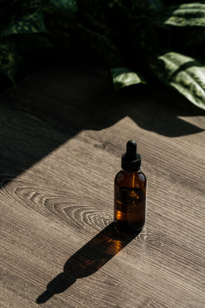 A brown glass dropper bottle sits on a wooden surface with light streaming in from the side. Leaves are partially visible in the background.