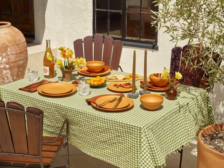 Outdoor dining table set with a green checkered tablecloth, terracotta plates, bowls, cutlery, glassware, candles, a bottle of wine, yellow flowers, and small tomatoes. Wooden chairs surround the table.