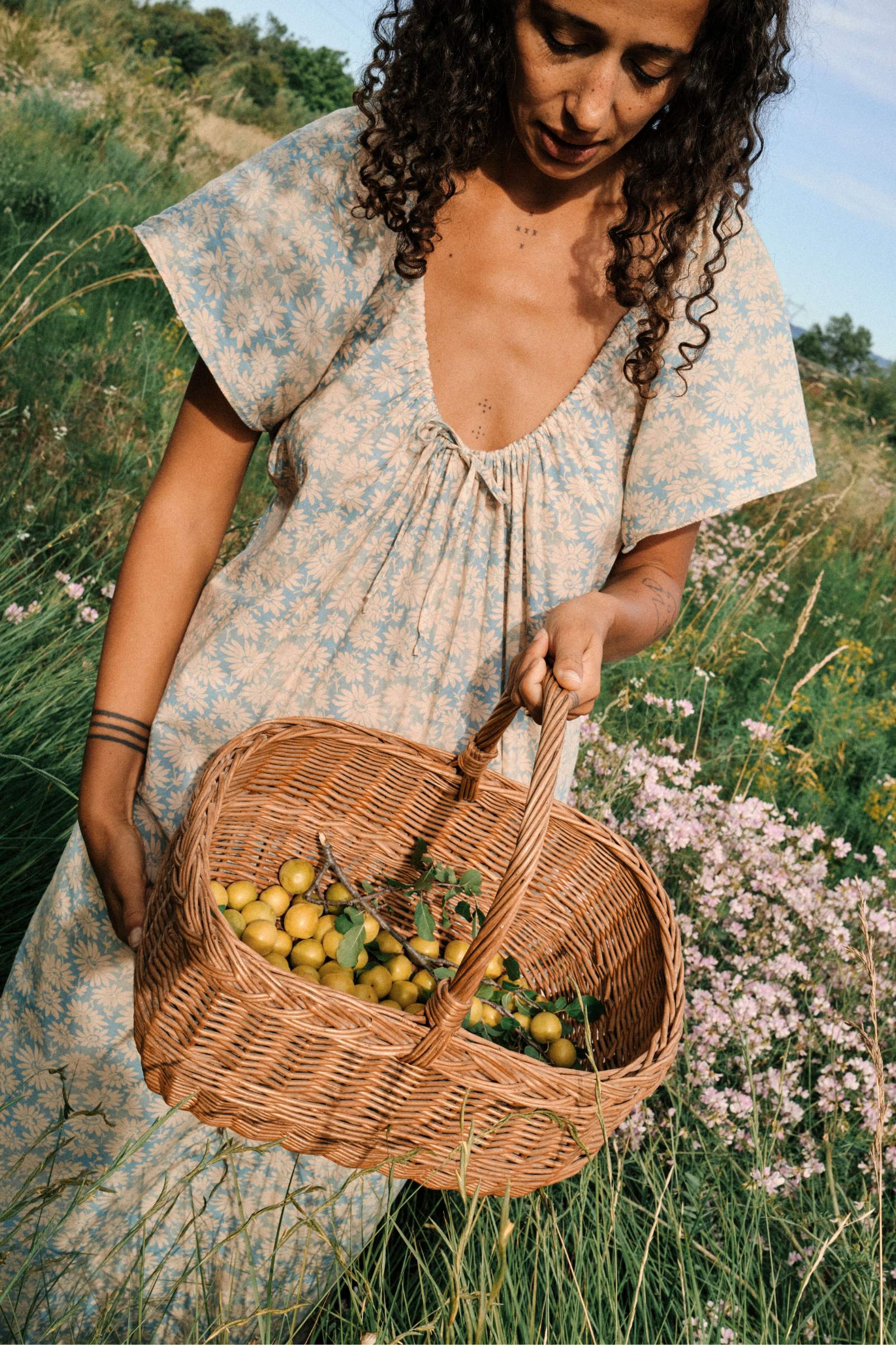 A woman in a floral dress stands in a meadow holding a wicker basket filled with yellow cherries.